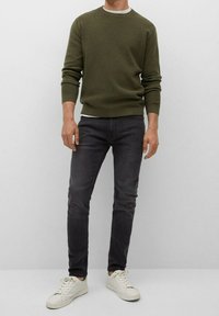 Muž v olivově zeleném svetru, černých slim-fit džínech a bílých teniskách stojí proti jednolitému světlému pozadí.