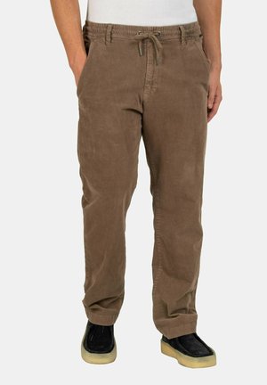 Braune Cordhosen mit elastischem Bund und Kordelzug, getragen mit schwarzen Schuhen mit hellen Sohlen, gezeigt von der Taille bis zu den Füßen.