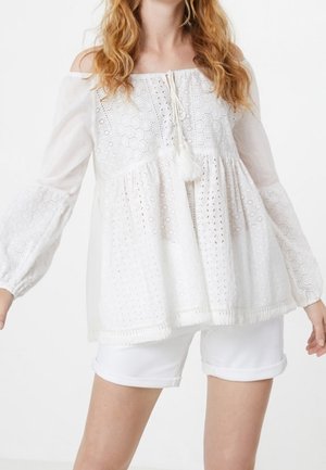 Femme portant un chemisier blanc à œillets épaules dénudées avec liens à pampilles et un short blanc avec ourlet roulotté sur un fond uni.