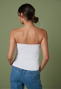 Haut blanc sans bretelles avec une texture lisse, associé à un pantalon en denim bleu. L'arrière-plan est d'un vert doux, mettant en valeur les couleurs des vêtements.