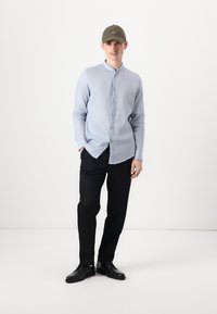 Lyseblå langermet skjorte med knapper, mørke bukser, svarte sko, og en olivenfarget caps. Slim fit med ståkrage og glatt tekstur.