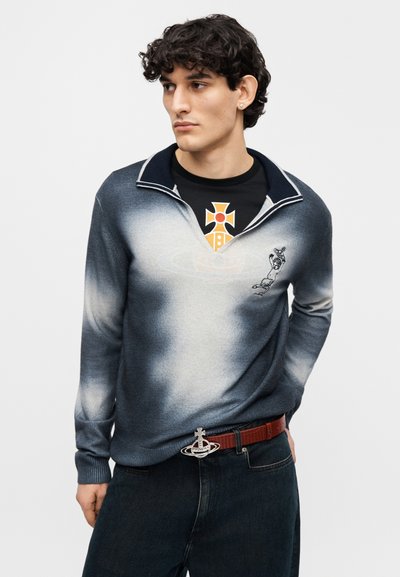 Πουλόβερ σε διαβαθμισμένη μπλε-γκρι πλέξη, με γιακά πόλο και κεντητό σχέδιο. Φορεμένο πάνω από μαύρο μπλουζάκι με πορτοκαλί γραφικό.