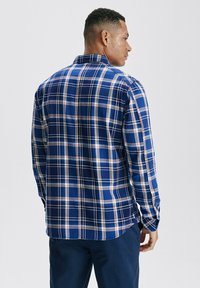 Niebieska koszula w kratę z długim rękawem, z kołnierzykiem zapinanym na guziki, z białymi i brązowymi akcentami oraz luźnym krojem. Pokazuje widok z tyłu oraz detale rękawów.