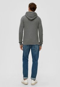 Man wearing grey hooded sweater, blue cuffed jeans, and white sneakers standing facing away against plain background.