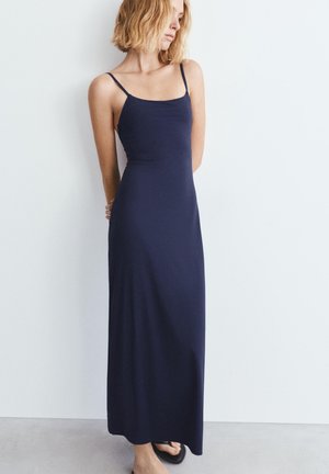 Žena s blond vlasmi v námornícky modrých bezrukávových maxi šatách a čiernych sandáloch stojaca s rukami za chrbtom pri bielej stene.