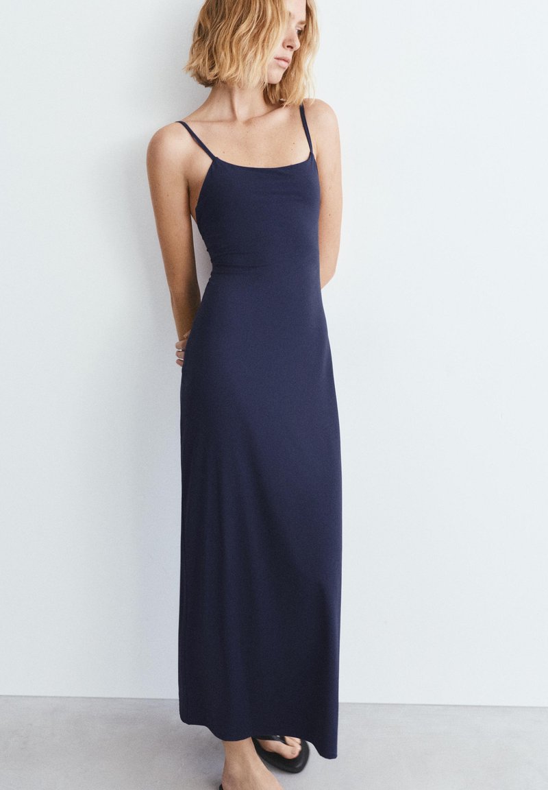 Femme aux cheveux blonds portant une robe longue sans manches bleu marine et des sandales noires, debout les mains derrière le dos contre un mur blanc.