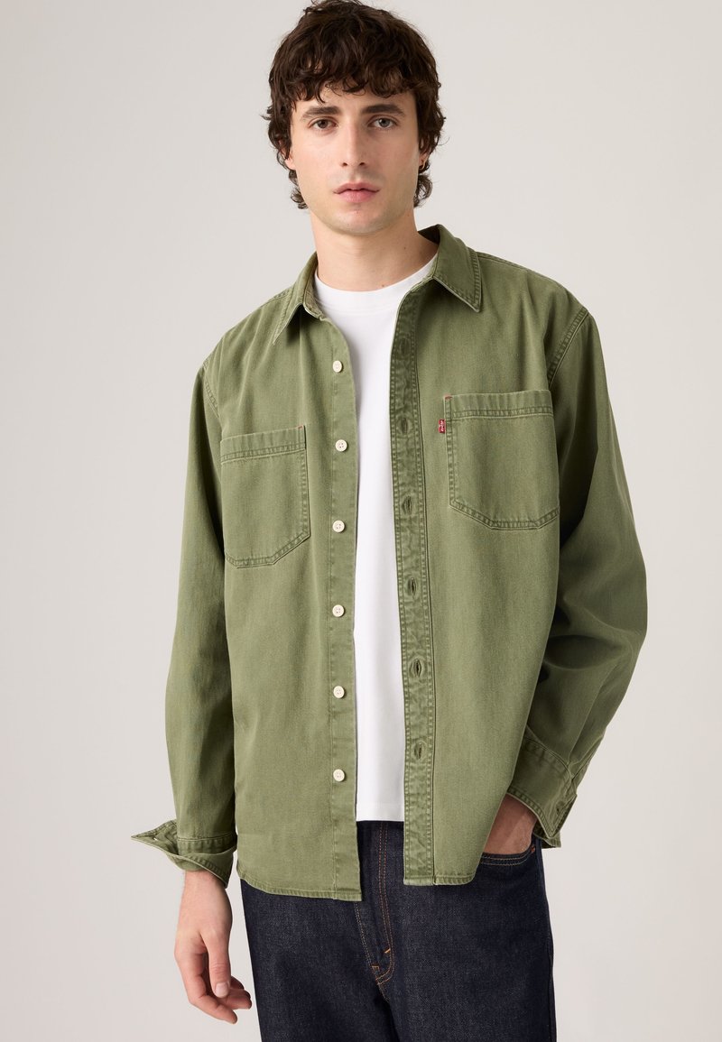 Giovane uomo con camicia verde oliva sbottonata sopra una t-shirt bianca e jeans blu scuro, in piedi davanti a uno sfondo chiaro uniforme.
