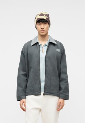 Giovane uomo con una giacca grigia con cerniera sopra una camicia azzurro chiaro, pantaloni bianchi e un berretto beige e marrone a motivo, su uno sfondo semplice.