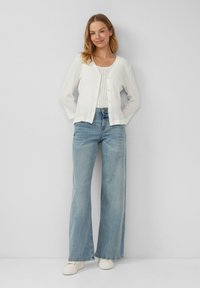 Witte geribde cardigan met knopen, gedragen over een witte tanktop. Gecombineerd met lichtblauwe wijde jeans en witte sneakers.