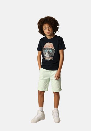 T-shirt graphique noir avec un design de crâne et de lunettes de soleil, associé à un short vert menthe et des baskets blanches. Cheveux bouclés, mains dans les poches.
