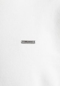 Petite étiquette métallique rectangulaire avec le mot "SMILODOX" attachée à un fond en tissu blanc uni.