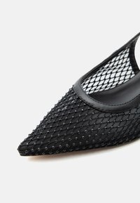 Μαύρο παπούτσι με μυτερή μύτη, με υφή από δικτυωτό ύφασμα που έχει μικρές ανυψωμένες κουκκίδες και δερμάτινη επένδυση.