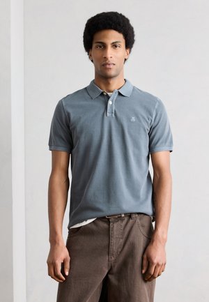 Man with dark curly hair wearing a blue short-sleeve polo shirt and brown pants, standing against a plain light background.