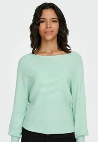 Mündiroheline soonikkoes kampsun laia kaelusega ja pikad kergelt täispuhutud varrukad, mis on kombineeritud mustade pükstega. Pehme tekstuur ja liibuv siluett.