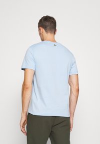 Camiseta de algodón azul claro con cuello redondo y mangas cortas. Logotipo verde notable de Lacoste en la parte posterior cerca del cuello.