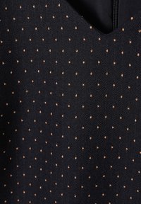Black fabric with a fine, consistent pattern of small beige dots. The material appears textured with a slight sheen, featuring a V-neck design.