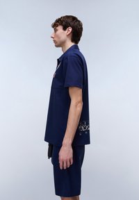 Ung mand stående i profil iført en mørkeblå polo med korte ærmer og matchende shorts mod en ensfarvet, lys baggrund.