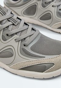 Gros plan de chaussures de sport grises en tissu mesh, avec surpiqûres synthétiques, accents en daim et semelles en caoutchouc texturé sur fond blanc.