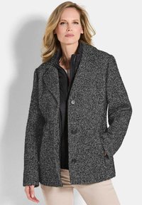 Blonde vrouw in een gestructureerde zwart-grijze knopenjas boven een zwart zipjack en beige broek, staand tegen een witte achtergrond.