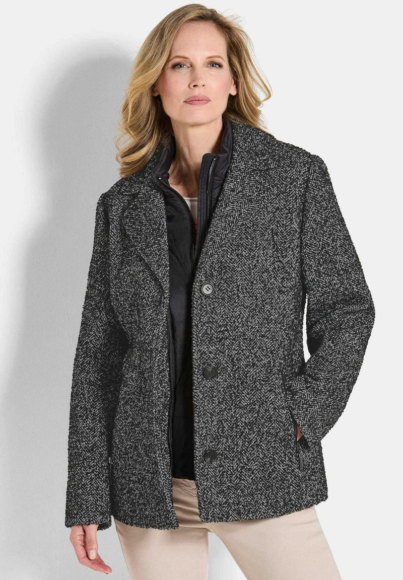 Blonde vrouw in een gestructureerde zwart-grijze knopenjas boven een zwart zipjack en beige broek, staand tegen een witte achtergrond.