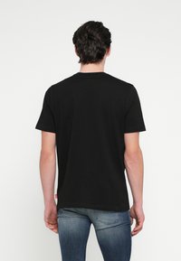 Čierne bavlnené tričko s klasickým okrúhlym výstrihom, krátkymi rukávmi a rovnakým lemom; na spodnej časti má malú červenú značkovú etiketu.