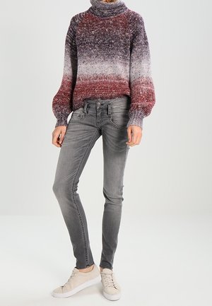 Person trägt einen gestrickten Rollkragenpullover mit Farbverlauf von Bordeaux zu Grau, graue Skinny-Jeans und beige Sneaker und steht vor einem schlichten Hintergrund.