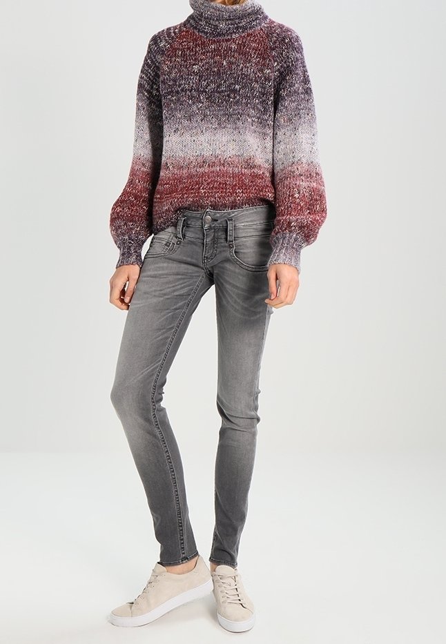 Personne portant un pull à col roulé en maille dégradée du bordeaux au gris, un jean skinny gris et des baskets beige, debout devant un fond uni.