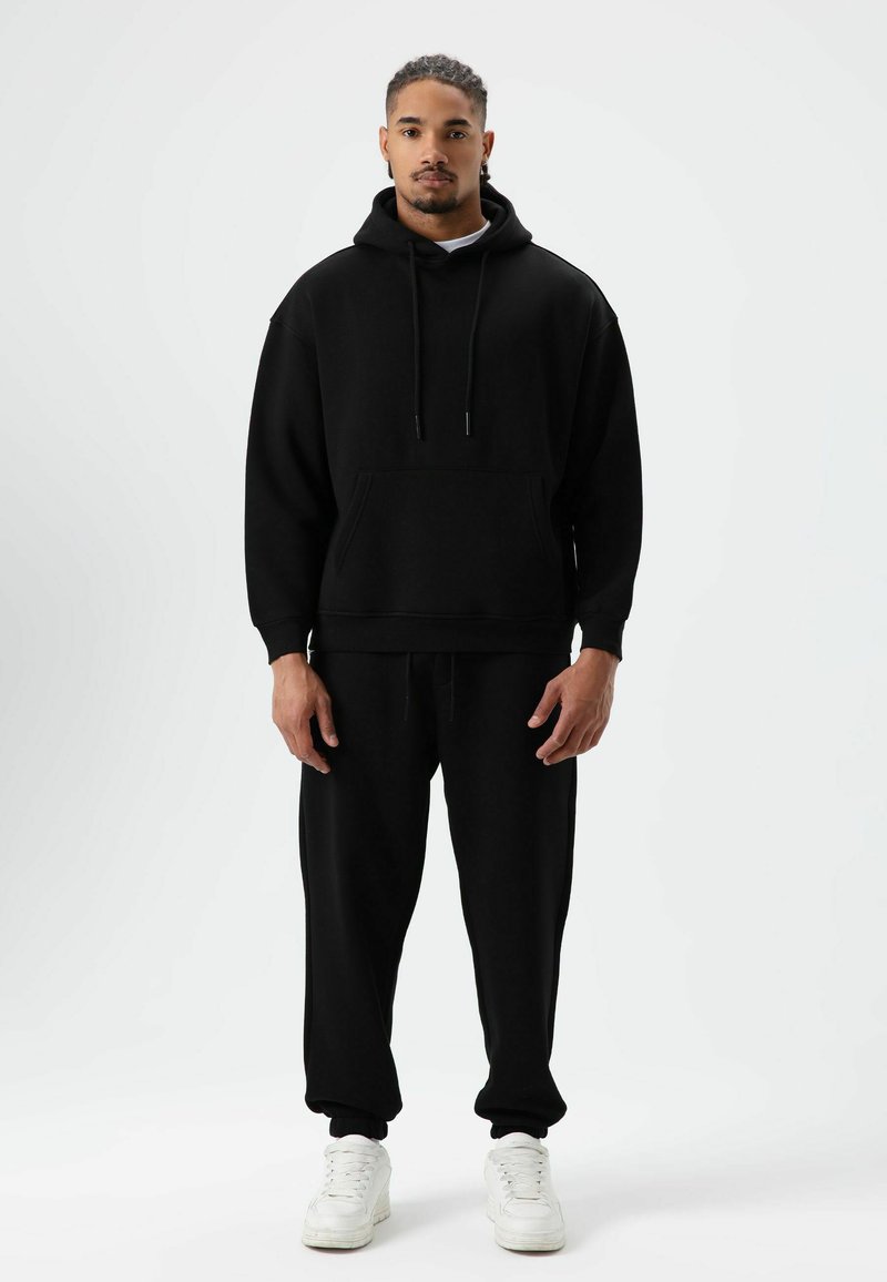 Zwarte katoenen hoodie met capuchon en trekkoord, en een voorkantzak, gecombineerd met bijpassende joggingbroek. Model draagt witte sneakers. Eenvoudig ontwerp, relaxte pasvorm.