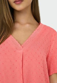 Koraalkleurige blouse met een V-hals, textuurstof met verhoogde stippen en kantaccenten langs de halslijn. Korte mouwen en een losse pasvorm.