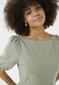 Olivno zelena obleka s kratkimi pufastimi rokavi, kvadratnim izrezom in subtilnimi šivi. Gladek material z mehko teksturo. Zlati uhani v obliki obroča.