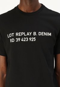 Svart bomull t-skjorte med hvit trykt tekst 'LOT REPLAY B. DENIM RD 39 423 925' i fet, grafisk stil over brystet.