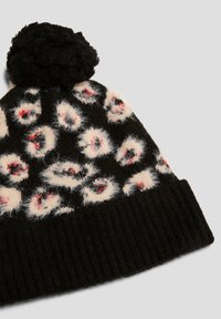 Zwarte gebreide beanie met witte pluizige luipaardvlekken en rode accenten, afgewerkt met een zwarte pompon. Heeft een geribbelde zwarte rand.