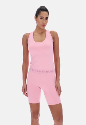 Kvinne iført en rosa ermeløs tanktopp og matchende rosa sykkelshorts med «Ted Baker Sport»-midjebånd, stående foran en ensfarget bakgrunn.