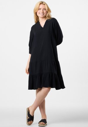 Czarna sukienka do kolan z fakturowanej tkaniny, z dekoltem w kształcie litery V, rękawami do trzech czwarte oraz falbaną u dołu. Nosi ją w czarnych sandałach.