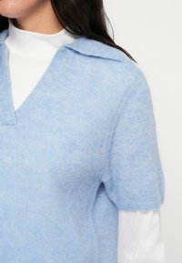 Γαλάζιο πουλόβερ με κοντά μανίκια, V-λαιμόκοψη και γιακά. Μια απαλή υφή, με εμφάνιση πολλαπλών στρωμάτων πάνω από λευκό τοπ με ψηλό γιακά.