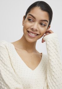 Pull en tricot crème avec un décolleté en V, motif en losanges texturé et poignets côtelés. Le matériau semble doux et léger.