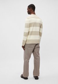 Leichter, gerippter Pullover in creme- und beige gestreift. Mit rundem Halsausschnitt und langen Ärmeln, kombiniert mit grauen Hosen und schwarzen Schuhen.