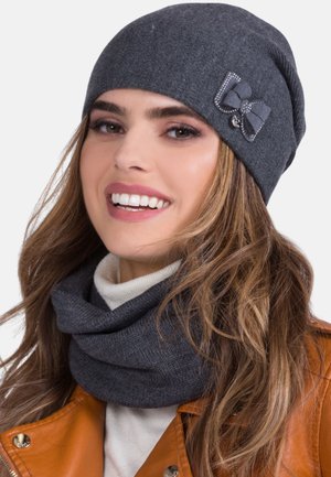 Woman smiling, wearing a dark gray knit beanie with a bow detail, matching neck warmer, white turtleneck, and brown leather jacket.