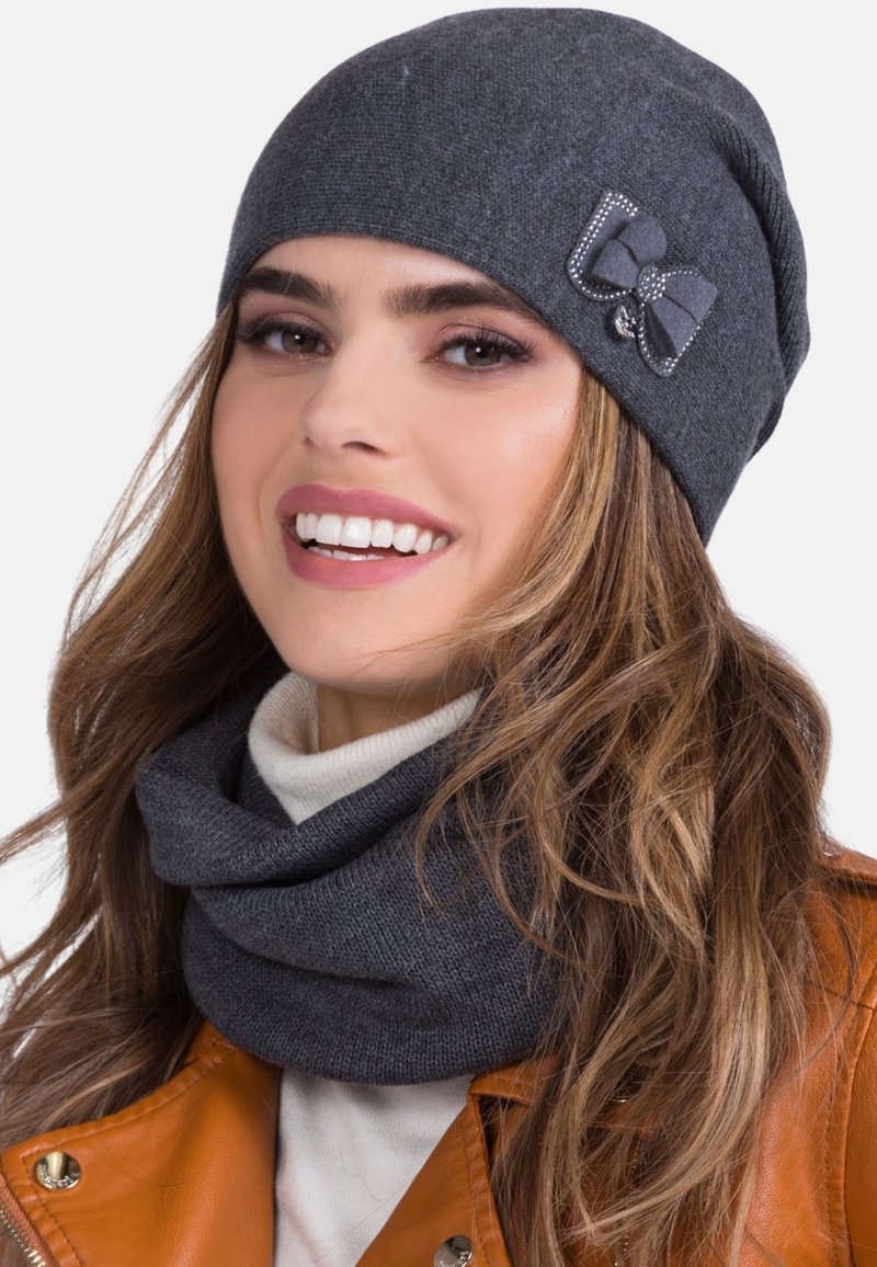 Woman smiling, wearing a dark gray knit beanie with a bow detail, matching neck warmer, white turtleneck, and brown leather jacket.
