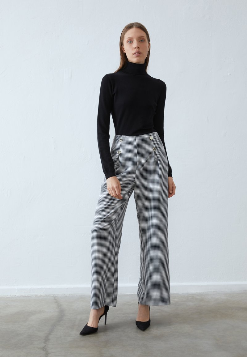 Maglione a collo alto nero abbinato a pantaloni a vita alta e gamba larga con motivo a scacchi, dotati di bottoni dorati e una texture di tessuto liscia.