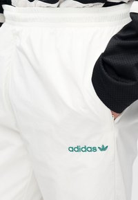 Λευκά ελαφριά joggers με ελαστική μέση, που διαθέτουν πράσινο κεντημένο λογότυπο "adidas" και διακοσμητικό φύλλο. Λείο υφή.