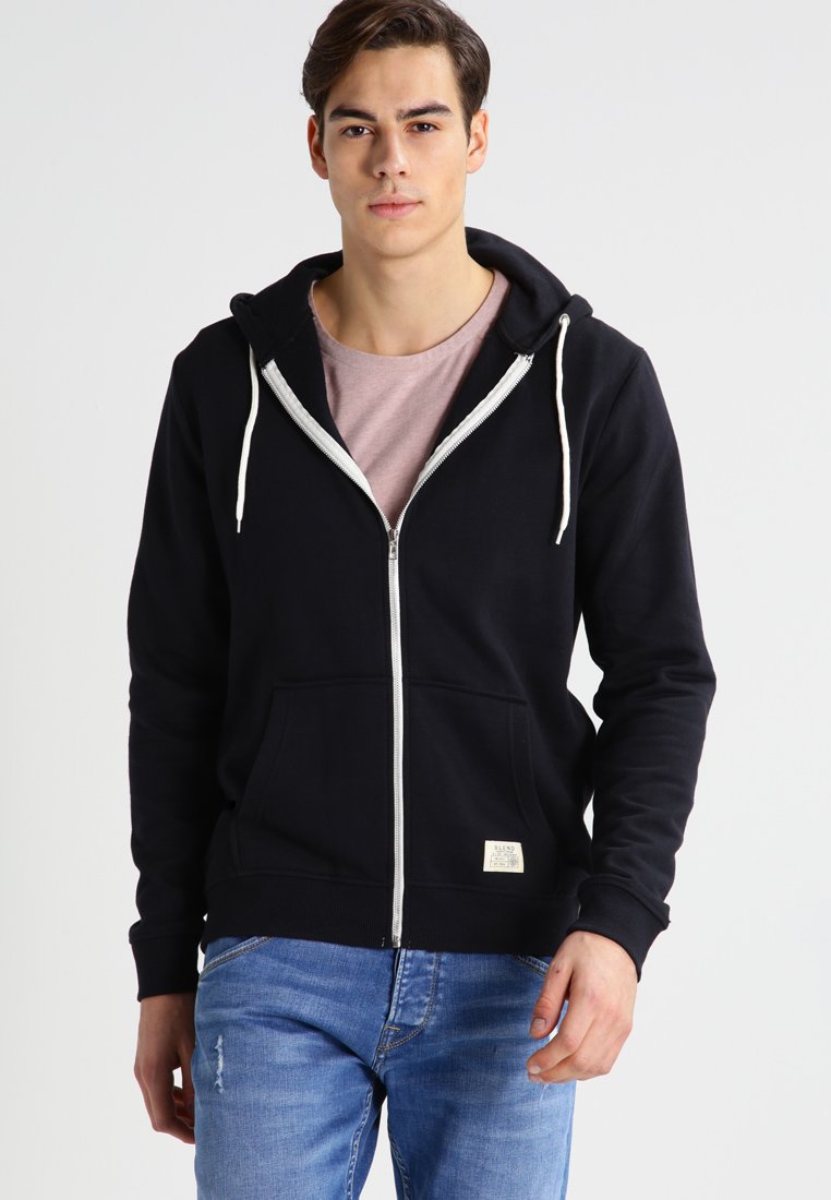 Sweat à capuche noir zippé avec une poche avant, cordons de serrage blancs et une petite étiquette. Porté sur un t-shirt rose clair et associé à un jean bleu.