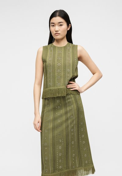 Γυναίκα με μακριά μαύρα μαλλιά που φοράει αμάνικη ελιά πράσινη πλεκτή μπλούζα και ταιριαστή φούστα με γεωμετρικά σχέδια και λεπτομέρειες με φουντάκια.