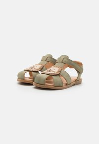 Grønne suede sandaler med åpne tær, med en tan løveansikt-akse på fremsiden og kryssende stropper for sikker passform. Flat såledesign.