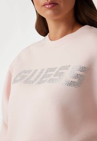 Ružičasti dukserica s okruglim izrezom, sadrži srebrni strazni logo "GUESS" na prednjem dijelu, s glatkom teksturom tkanine.