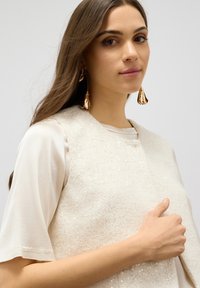 Top senza maniche color crema con paillettes sovrapposto a una camicia leggera color beige. Presenta orecchini pendenti che assomigliano a petali di fiore.
