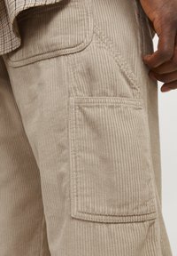 Beige corduroy cargo pants with vertical ribbed texture, side pocket, and detailed stitching accents. Close-up view highlights fabric quality.