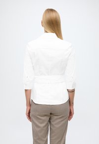 Camicia bianca aderente con maniche a tre quarti, vita con cintura e dettaglio sul dorso; abbinata a pantaloni a quadretti marroni e beige.