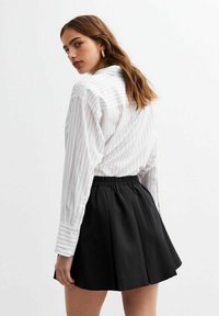 Chemise blanche à rayures verticales avec des manches retroussées, associée à une jupe skater noire avec une taille élastique, vue de dos.