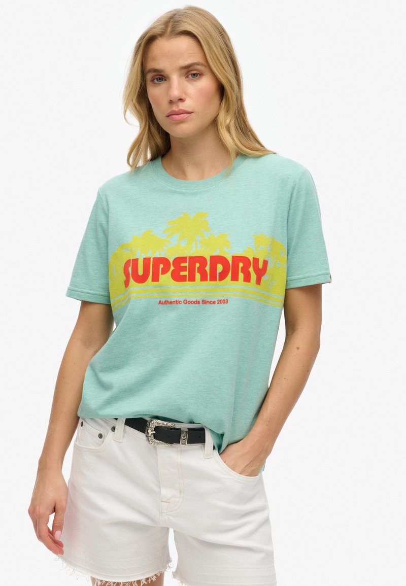 Lys teal bomulds t-shirt med et iøjnefaldende rødt "SUPERDRY" logo, palmetræ grafik og korte ærmer. Parret med hvide shorts og et sort bælte.